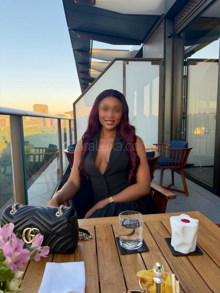 Zara Luxa wearing a black sleeveless vest-style dress sits at an outdoor rooftop restaurant table, with a black handbag, glass of water, flowers, and a city view in the background.