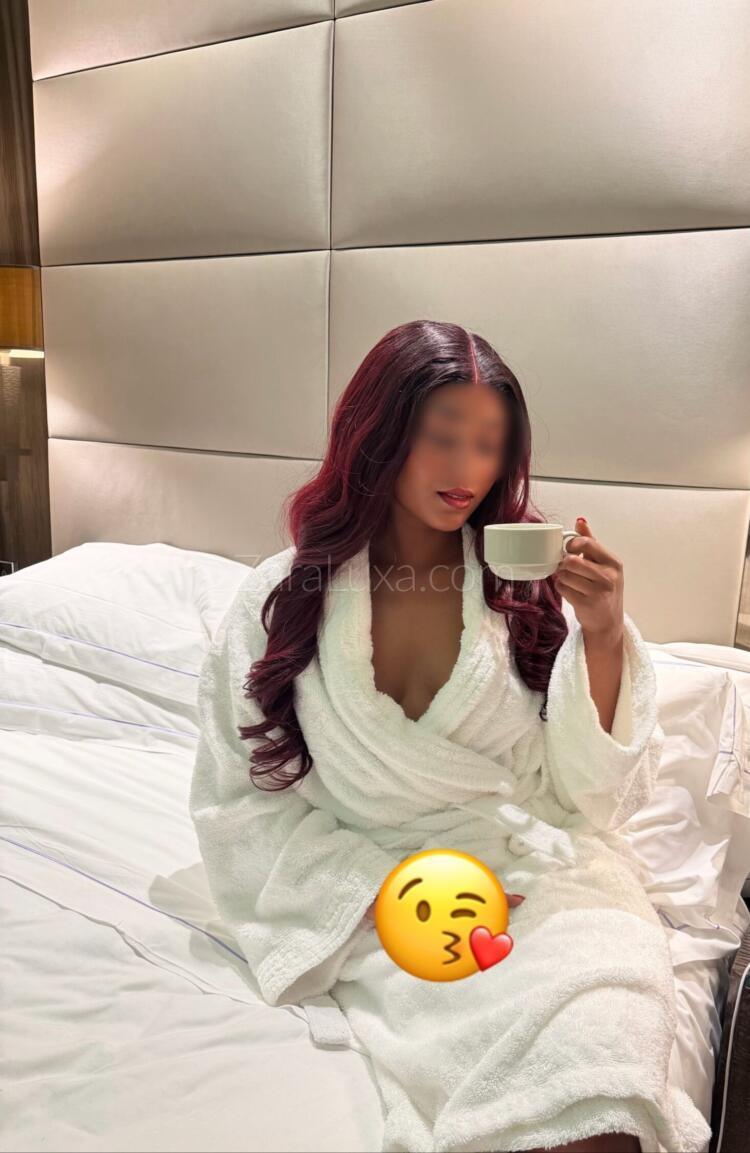 Zara Luxa in a white bathrobe with a cup of tea, on a bed in a hotel bedroom