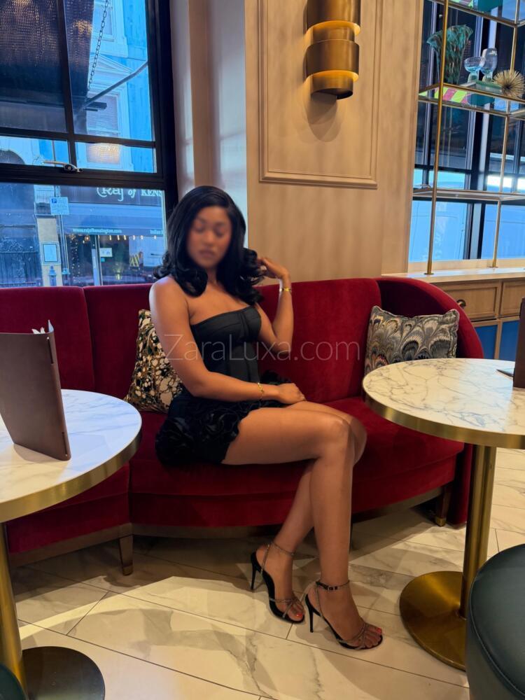 Zara Luxa in a black dress and heels, seated on a red velvet sofa in a Kensington hotel cocktail lounge