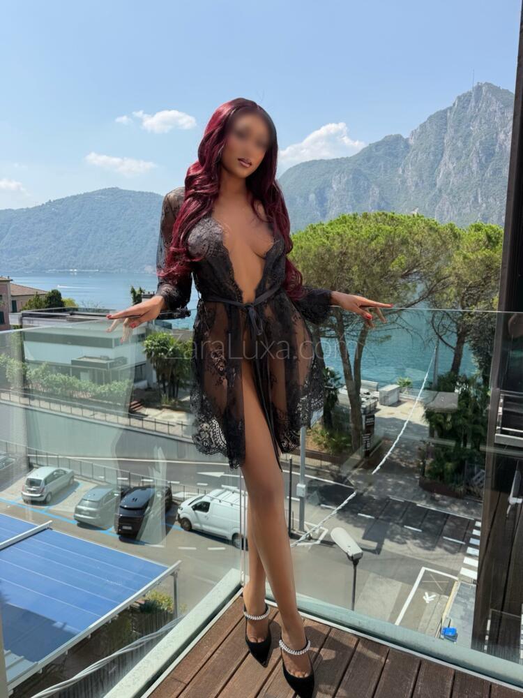 Zara Luxa with long burgundy hair, in a black lace robe and heels, on a balcony overlooking Lake Como